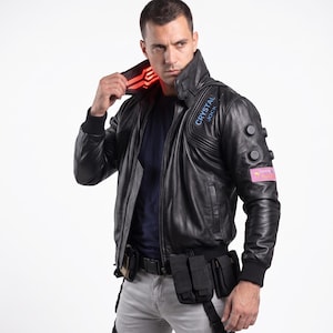Pode incluir: Um blusão bomber de couro preto com gola alta e painel de luz vermelha, punhos canelados e um emblema bordado em azul com o texto CRYSTAL JOCK. A manga esquerda inclui três botões pretos circulares e um emblema retangular cor-de-rosa.