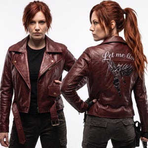 May include: A dark red leather motorcycle jacket featuring a wide notched collar, a front zipper, and a belted waist. The back displays a graphic of a winged figure with the script text Let me live, showcasing a structured fit and metal hardware.