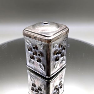 Pode incluir: Um pequeno recipiente de metal prateado com uma parte superior quadrada e um corpo cilíndrico. O corpo é decorado com padrões florais e pequenos orifícios circulares. O recipiente é refletido em uma superfície brilhante.