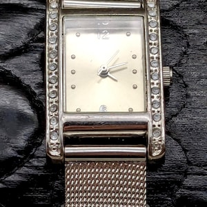 May include: Silver rectangular wristwatch with a mesh strap and a light-coloured face. The watch has a silver bezel with small, clear stones. The watch face has small, round markers and the number 12 at the top.