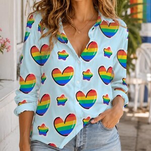 May include: A light blue button-down shirt with a repeating pattern of rainbow-colored hearts and star shapes. The shirt has long sleeves and is worn with denim shorts. The overall theme is pride and inclusivity.
