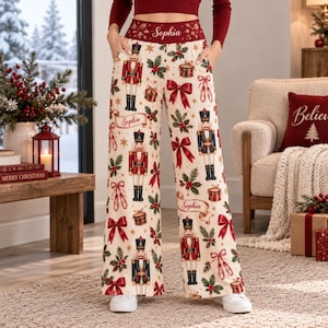 Puede incluir: Pantalones de descanso de pierna ancha en color crema con un estampado navideño festivo de cascanueces, lazos rojos, hojas de acebo y cajas de regalo. La cintura es de color rojo oscuro con el nombre Sophia en blanco, también en cintas.