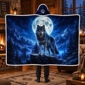 May include: A hooded blanket featuring a dark blue night scene with a full moon, a wolf standing on a rocky outcrop, and a forest of pine trees. The hood displays a white paw print graphic. The entire blanket has a white trim border.