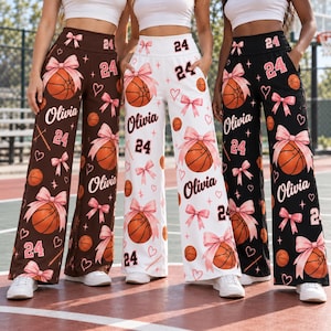 May include: High-waisted wide-leg pants in brown, white, and black, featuring a repeating pattern of basketballs tied with pink bows, the name Olivia, and the number 24. The pants have an elastic waistband and side pockets.