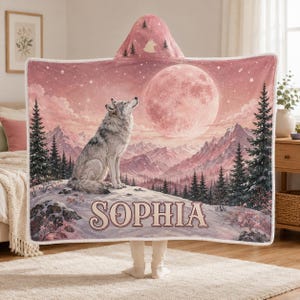 May include: A hooded blanket featuring a pink-toned landscape with a wolf sitting on a snowy hill under a large full moon. The scene includes pine trees and mountains. The name SOPHIA is printed in large, dark letters across the lower section.