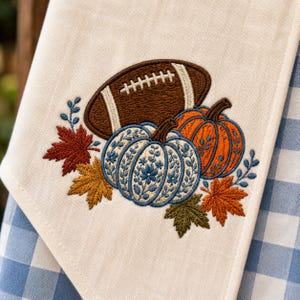 May include: A cream-colored fabric towel featuring an embroidered design of a brown football, an orange pumpkin, and a white pumpkin with blue floral patterns. The arrangement is accented with red, orange, and green embroidered autumn leaves.