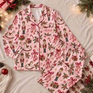 May include: A light pink long-sleeve pajama set with a button-down top and matching pants. The fabric features a repeating pattern of nutcrackers, rocking horses, gift boxes, ballet slippers, and candy canes. The pocket has the name OLIVIA.
