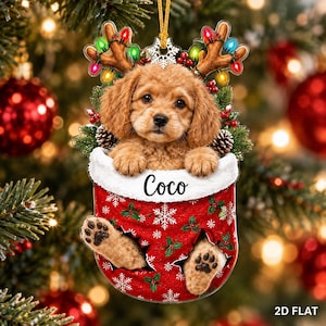 May include: A flat Christmas ornament featuring a light brown puppy peeking out of a red stocking with white snowflake and holly patterns. The puppy wears reindeer antlers with colorful lights. The name Coco is printed on the white cuff.