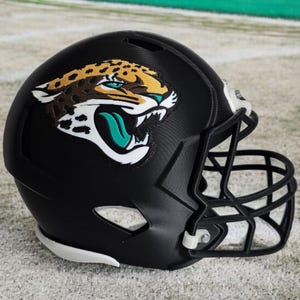 May include: A black football helmet featuring the Jacksonville Jaguars logo. The logo depicts a jaguar's head with teal, gold, and white accents. The helmet has a black facemask and white padding.