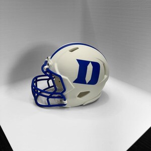 May include: A white football helmet featuring a blue face mask and a blue letter D logo on the side. The helmet includes ventilation slots along the top and sides, with a blue stripe running down the center from the front to the back of the shell.