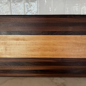Puede incluir: Una tabla de cortar rectangular de madera que presenta una franja horizontal central ancha de color claro, flanqueada por dos franjas horizontales más oscuras de color marrón intenso en la parte superior e inferior, con un acabado pulido.