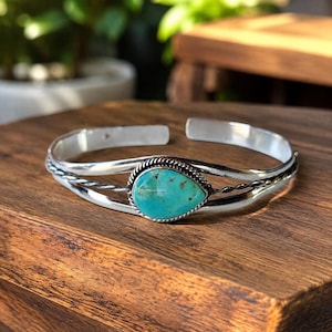 May include: A silver cuff bracelet featuring a central teardrop-shaped turquoise stone. The band is constructed from two smooth silver wires and one twisted silver wire that frames the stone, creating a detailed open-ended metal design.