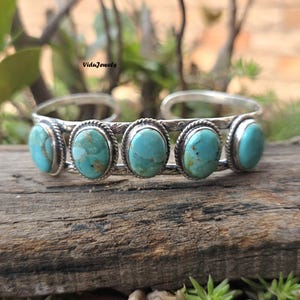 May include: A silver cuff bracelet featuring five oval-shaped turquoise stones set in rope-textured bezels. The band has an oxidised finish with etched detailing between the stones. The text ViduJewels is visible above the bracelet.