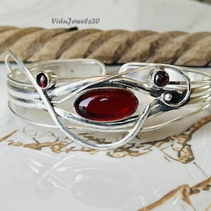 May include: A silver cuff bracelet featuring a central oval, deep red gemstone and smaller red gemstones. The bracelet has an open design with curved silver detailing.