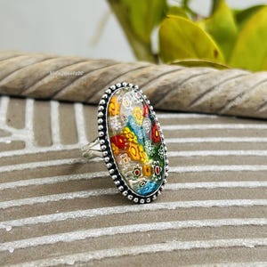 May include: An oval-shaped ring featuring a millefiori glass cabochon with a vibrant pattern of small red, yellow, blue, and white flower-like shapes set against a clear background, encased in a silver-toned metal frame with a beaded edge.