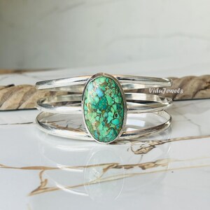 Può includere: Un bracciale a polsino in argento con una grande pietra ovale di turchese nei toni del verde e dell'oro. Il bracciale ha tre fasce parallele e una finitura lucida. Il gioiello è esposto su una superficie di marmo.
