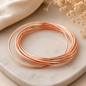 Puede incluir: Una colección de finas pulseras de metal color cobre, apiladas y ligeramente superpuestas. Las pulseras están dispuestas sobre una superficie clara y texturizada, con una tela suave de tono neutro en el fondo. El texto "Vide Jewels" es visible.