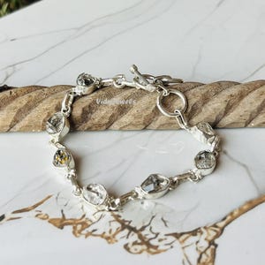 Puede incluir: Una pulsera de plata con piedras preciosas claras de forma irregular. La pulsera tiene un cierre de palanca y se muestra sobre una superficie de madera sobre un fondo de mármol. Las piedras están engastadas en eslabones de plata.