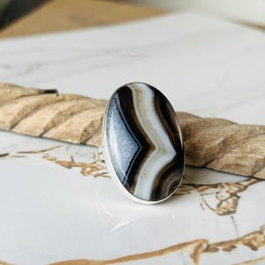 May include: A silver ring featuring a large, oval-shaped banded agate gemstone. The stone displays prominent, smooth layers of black, white, and translucent grey in a chevron-like pattern, securely set within a polished silver metal bezel setting.