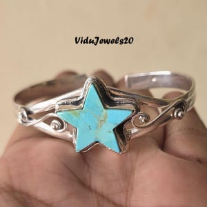 Peut inclure: Un bracelet manchette en argent orné d'une pierre turquoise en forme d'étoile avec des veines brunes naturelles. La bande métallique présente des détails en filigrane tourbillonnant de chaque côté de l'étoile. Le texte ViduJewels20 est visible.