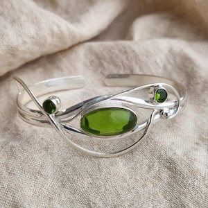 May include: A silver cuff bracelet featuring a large, oval-shaped, translucent olive-green stone at the centre, accented by two smaller, round, vibrant green stones set within a polished, flowing, multi-strand metal wire design.