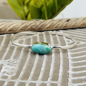 May include: A silver cuff bracelet featuring a single oval-shaped turquoise stone at the center. The polished silver band is open-ended, and the turquoise stone displays natural blue and green veining throughout its smooth, rounded surface.