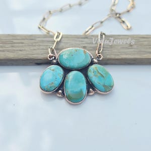 May include: A turquoise stone necklace with a silver chain. The pendant features four oval-shaped turquoise stones and one larger oval stone in the center. The necklace is displayed on a wooden plank. The text "Vida Jewels" is visible.