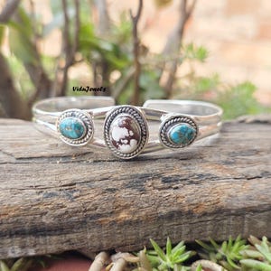 May include: A silver cuff bracelet featuring three oval stones set in twisted metal frames. The center stone is white with brown matrix patterns, while the two flanking stones are blue with dark brown veining. The text ViduJewels is visible.