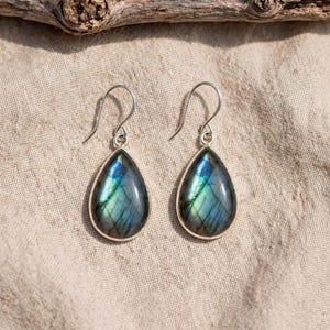 May include: A pair of teardrop-shaped labradorite earrings set in silver-toned metal. Each earring features a polished, iridescent stone displaying flashes of blue and green, suspended from a simple curved silver-toned ear wire hook.