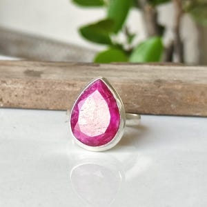 Peut inclure: Une bague en argent ornée d'une grande pierre précieuse rose vif en forme de poire, taillée à facettes et sertie dans un chaton en métal lisse. La pierre présente une teinte magenta vibrante avec des inclusions internes visibles.