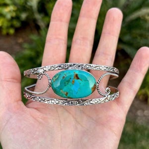 Könnte beinhalten: Ein silbernes Armband mit einem großen, ovalen Türkisstein mit braunen Matrixmustern. Das Metallband ist mit aufwendigen Blumengravuren und gedrehten Kordeldetails an den Rändern verziert, die den zentralen Edelstein kunstvoll einrahmen.
