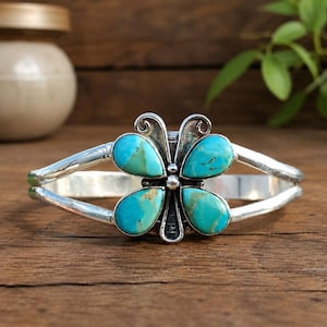 May include: A silver cuff bracelet featuring a central butterfly-shaped design. The wings are composed of four teardrop-shaped turquoise stones with brown matrix veining. The metal band is smooth and polished, extending from the central ornament.