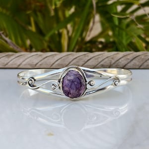 Puede incluir: Un brazalete de plata con una piedra preciosa central de color púrpura intenso, de corte rugoso y engastada en un bisel de metal pulido. La banda está formada por dos hilos de plata paralelos que se curvan en volutas decorativas.