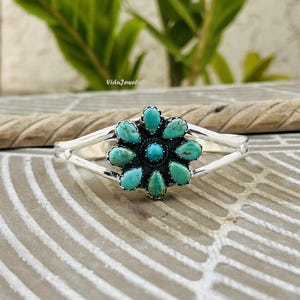 May include: A silver cuff bracelet featuring a central flower-shaped cluster of nine teardrop-shaped turquoise stones surrounding a small round turquoise centre. The metal band splits into two arms on each side of the floral centrepiece.
