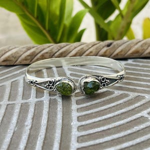 May include: A silver cuff bracelet featuring two raw, faceted green gemstones set at each open end. The metal band displays intricate filigree detailing and decorative scrollwork surrounding the stones, creating a balanced and symmetrical design.