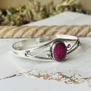 Puede incluir: Una pulsera de plata con una gema ovalada de color rojo intenso en el centro. La pulsera tiene un diseño abierto con remolinos decorativos y pequeñas cuentas de plata. Fondo de mármol.
