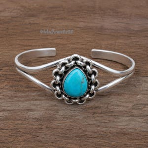 May include: A silver cuff bracelet featuring a central teardrop-shaped turquoise stone surrounded by a scalloped metal frame. The band consists of two smooth silver wires that meet at the decorative centre. The text ViduJewels20 is visible.