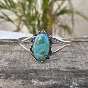 May include: A silver cuff bracelet featuring a large, oval-shaped turquoise stone with brown matrix veining. The stone is set in a decorative silver bezel with beaded and twisted wire detailing, attached to a split-shank silver band design.