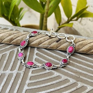 Könnte beinhalten: Ein silbernes Kettenarmband mit sechs ovalen, facettierten magentafarbenen Steinen in gedrehten Metallfassungen. Das Armband verfügt über einen Knebelverschluss mit einem runden Ring und einem T-Stab, die eine detaillierte Metallstruktur aufweisen.