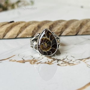 Può includere: Un anello in argento con un fossile di ammonite a forma di goccia come elemento centrale, caratterizzato da motivi a spirale marroni e beige. La fascia in metallo ha un design a più fili con piccole sfere d'argento ai lati. Testo "WiduJewels20".