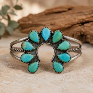 May include: A silver cuff bracelet featuring a horseshoe-shaped centerpiece adorned with nine teardrop-shaped turquoise stones. The metal band has a twisted rope texture, and the silver displays a darkened, oxidized finish around the stones.