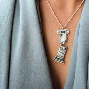 May include: A silver chain necklace with a broken column pendant. The pendant is made of white ceramic and has a black outline. The necklace is worn by a person wearing a light blue shirt.