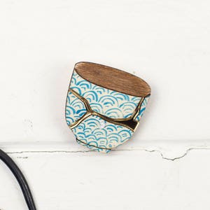 May include: A decorative wooden and ceramic piece with a Japanese Kintsugi design. The top is wood, the body is white with a blue wave pattern, and the cracks are filled with gold. Black metal tools are in the lower left corner.