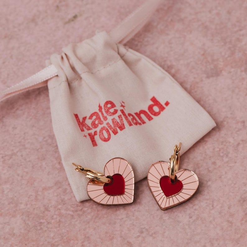 May include: A pair of gold hoop earrings with pink and red heart charms. The charms have a sunburst design and the text "kate + rowland" is printed on a white fabric bag.
