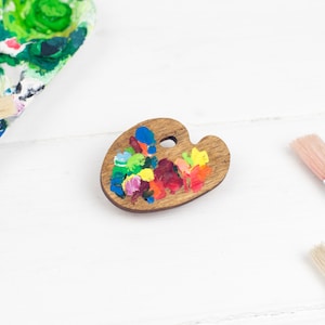 Könnte beinhalten: Eine Holzpalette für Künstler mit bunten Farbflecken. Die Palette ist tropfenförmig und hat ein Loch in der Mitte. Zwei Pinsel sind ebenfalls auf dem Bild zu sehen.