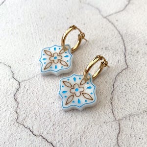 Puede incluir: Un par de pendientes de aro dorados con colgantes estampados en blanco, azul y marrón. Los colgantes tienen un diseño floral con detalles azules y contornos marrones. Los pendientes se muestran sobre una superficie blanca texturizada.