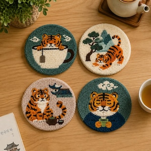 May include: Four circular tufted coasters featuring tiger illustrations. One shows a tiger in a teacup, another a tiger near a tree, a third a tiger with a teapot, and the fourth a tiger holding a cup. The designs use orange, white, green, and blue yarn.