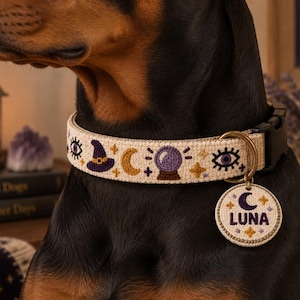 May include: A beige dog collar featuring embroidered purple witch hats, gold crescent moons, purple crystal balls, and eyes. A round, matching beige tag hangs from a brass D-ring, displaying a purple crescent moon, gold stars, and the name LUNA.
