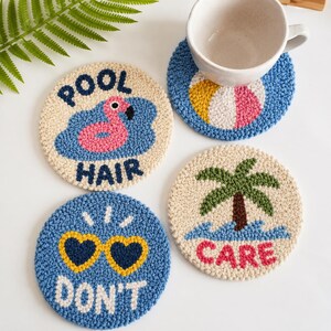 May include: Three circular punch needle coasters with textured yarn designs. One features a pink flamingo in blue water with the text POOL HAIR. Another shows a beach ball with a white mug on top. The third displays a palm tree and the text CARE.
