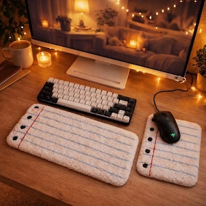 Puede incluir: Una configuración de escritorio de computadora con un teclado blanco y negro, un ratón negro y una alfombrilla de ratón estilo bloc de notas. La alfombrilla tiene líneas azules y un margen rojo. El escritorio es de madera.